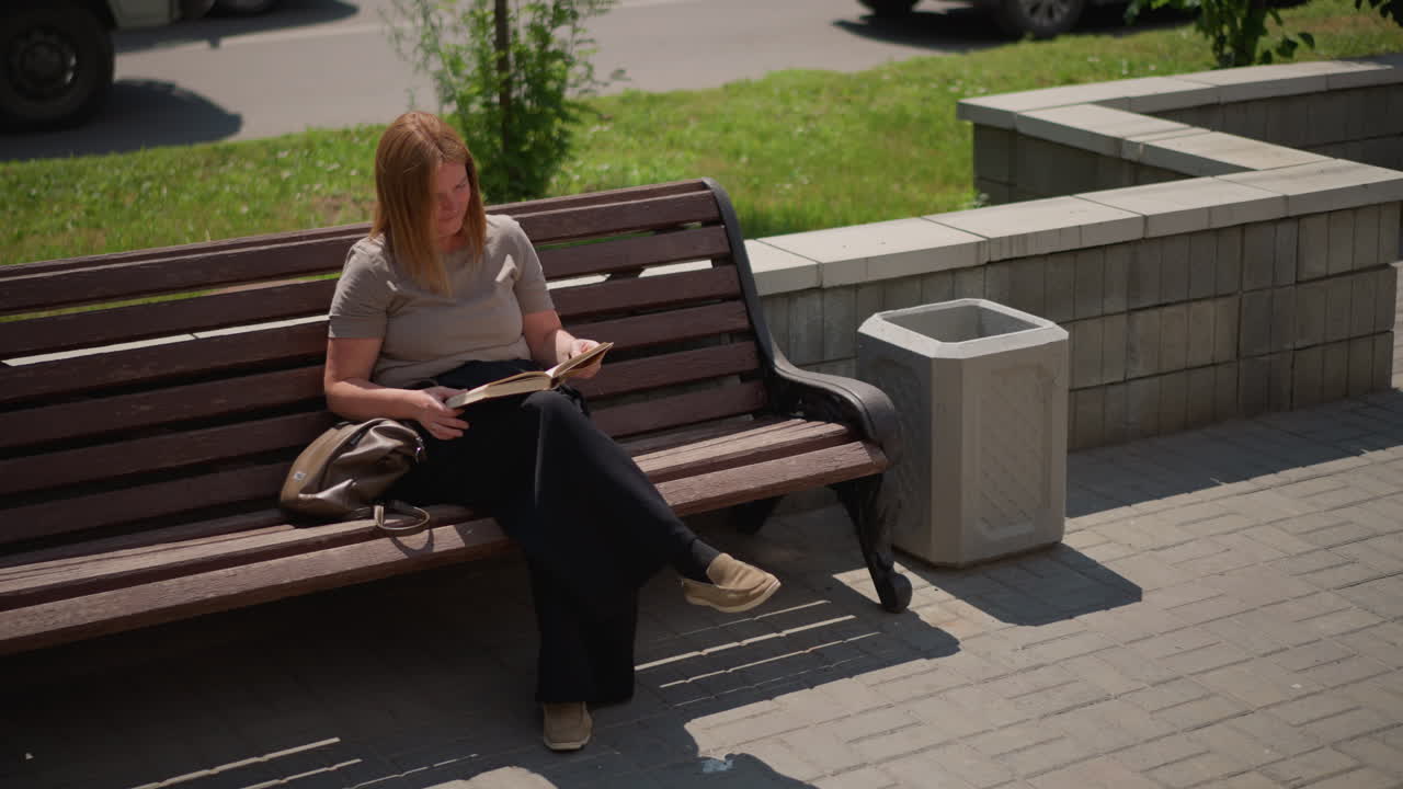 Pupil sitting on wooden bench reading and flipping page of book under bright sunlight, focused expression showing curiosity and calm, handbag beside, background with soft blur of grass