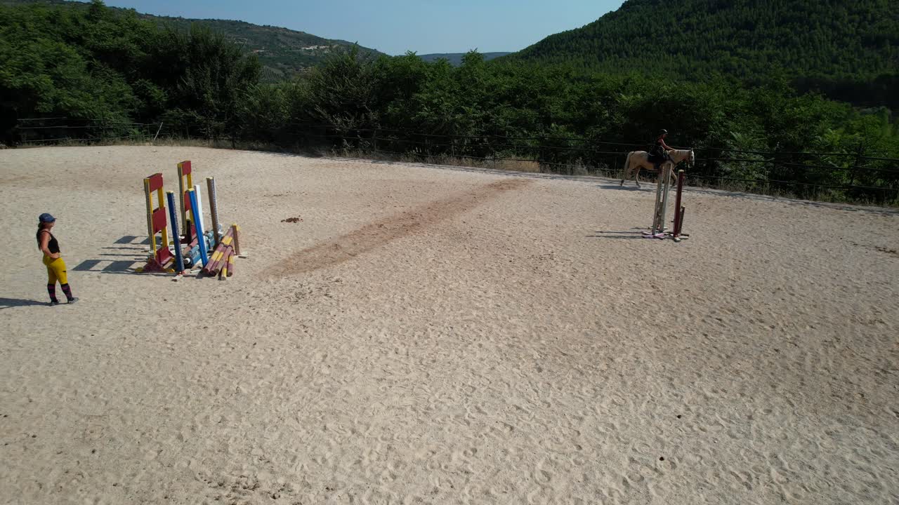 Pullback drone shot reveals a trainer in the middle of the arena teaching two women how to ride a horse