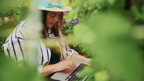 woman in straw hat working on laptop surrounded by vibrant flowers in a lush garden setting