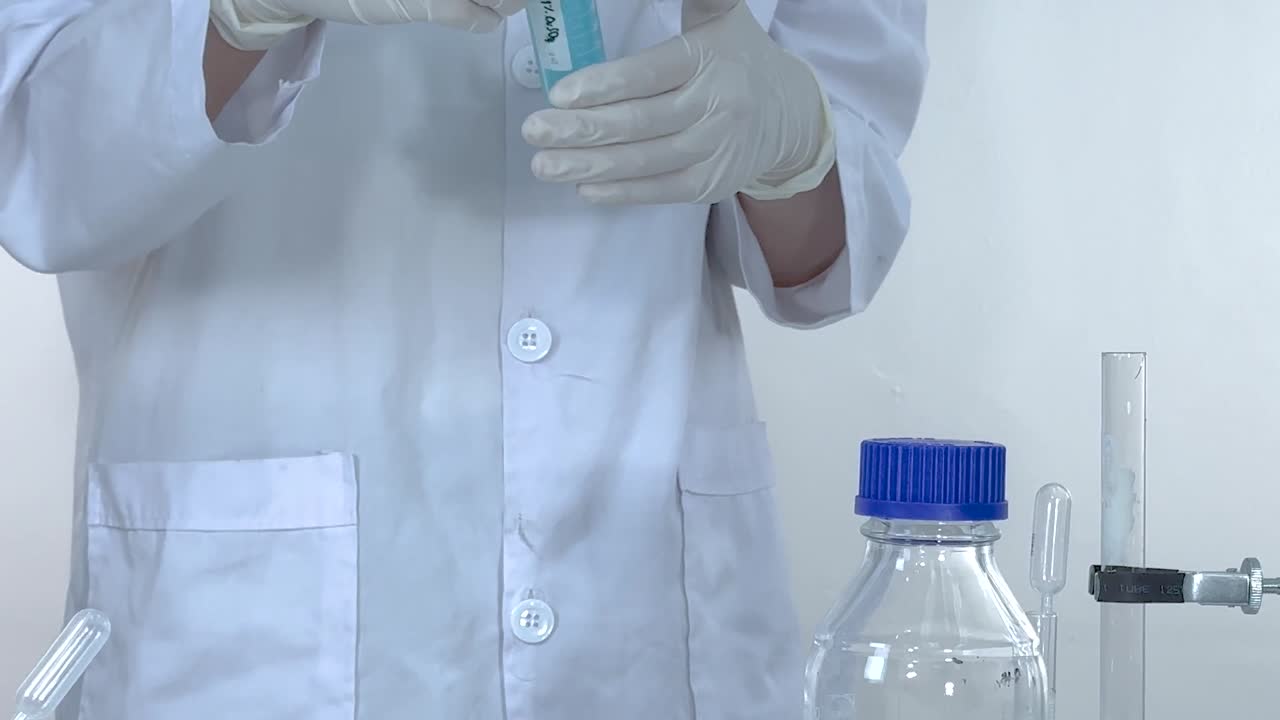 A lab technician carefully handles a blue liquid in a test tube using gloves.