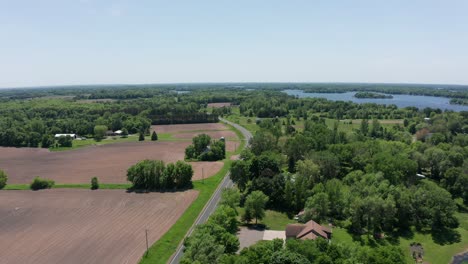 Tiro-Aéreo-Descendente-Volando-Sobre-Un-Camino-Rural-De-Tierras-De-Cultivo-A-Lo-Largo-De-La-Orilla-Del-Lago-En-Minnesota