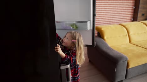 una niña linda abre la puerta del refrigerador alcanzando algo