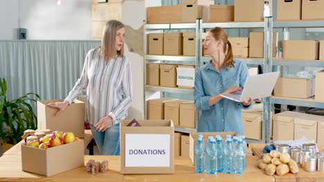 caucasian young female volunteer typing on laptop while her coworker packing donation boxes in charity warehouse