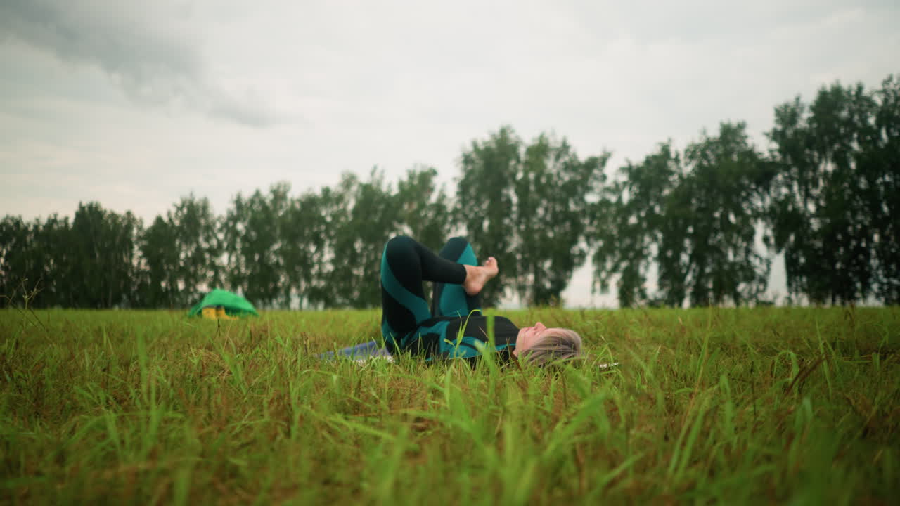 요가 매트 위에 누워있는 여성은 허리를 지지하는 손으로 브리지 포즈를 연습하고, 구름이 가득한 하늘 아래 광활한 풀에서, 멀리서 나무와 녹색 천을 가진 노란색 부츠를 배경으로합니다.
