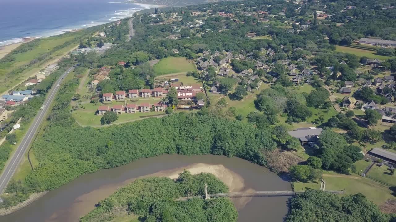 A Drone flying above and over a resort with rondavels in Hibberdene durban beach with a canal and river lagoon below and cars over a highway.