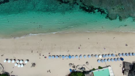 Imagen-Aérea-De-La-Playa-De-Melasti-En-Uluwatu-Bali,-Con-Varios-Hoteles-Y-Sombrillas-Visibles,-Así-Como-Olas-Que-Se-Acercan-A-La-Playa-Y-Gente-Paseando-En-La-Arena-Fue-Capturada-Por-Un-Dron