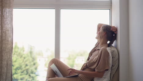 Young-woman-studies-via-laptop-sitting-on-windowsill-at-home