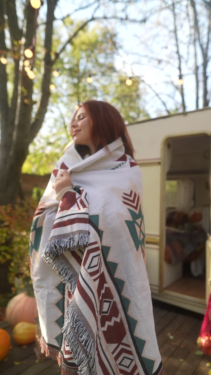 mujer envuelta en una manta por un remolque de campamento en otoño