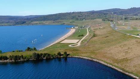 playa del lago con personas tomando el sol y bañándose, pequeños veleros navegando, jardín y área para caminar