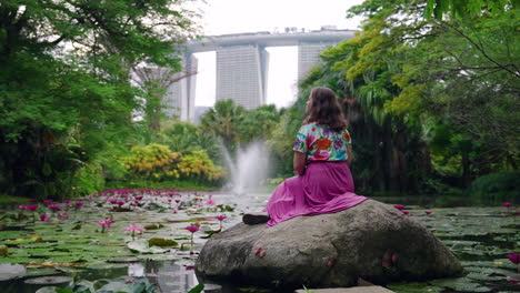 a lady in pink seated on the rock over pond filled with lilies at gardens by the bay with marina bay sands view in singapore