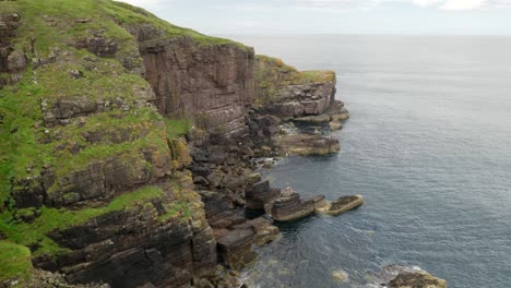 Looking-out-to-the-vast,-blue-Atlantic-Ocean-as-waves-gently-lap-against-a-tall-sea-cliff-in-the-ocean-while-seabirds-fly-around-the-grassy-clifftops