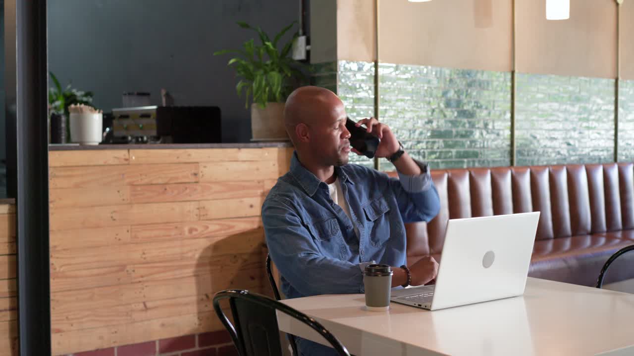 African American man in denim answering ringing smartphone for work smiling resuming laptop typing