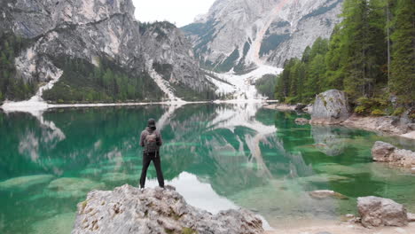 Backwards-revealing-drone-shot-of-a-male-model-standing-on-a-rock-overlooking-lake-Braies-in-the-Dolomites,-Italy