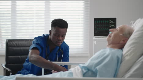 a black cardiologist doctor is talking to a 60-70-year-old patient lying on a bed in a hospital. a neurologist is talking to a patient. the patient is connected to an oxygen mask and an ecg device