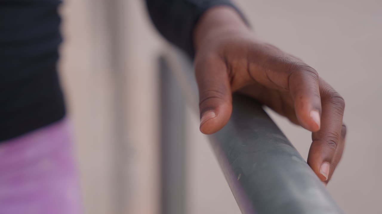 Primo piano di una mano su una ringhiera di metallo che mostra riscaldamento e concentrazione, le dita si aggrappano delicatamente alla sbarra, leggings viola e top nero leggermente visibili, lieve sudore sulla pelle, profondità di campo ridotta, ambiente urbano tranquillo.