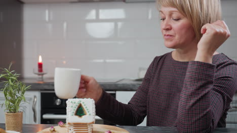 mujer disfruta de una bebida caliente, descansando la cabeza sobre su mano mientras mira contemplativamente a la casa de pan de jengibre en una bandeja de madera, luz de vela cálida y decoración de cocina rústica