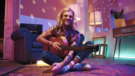 young stylish caucasian man with long hair sitting on the floor and playing the guitar in a retro party at home