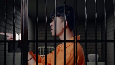 vista lateral de cerca de un prisionero masculino asiático con esposas de pie en la cárcel, señalando con el dedo, hablando en contra y culpándote
