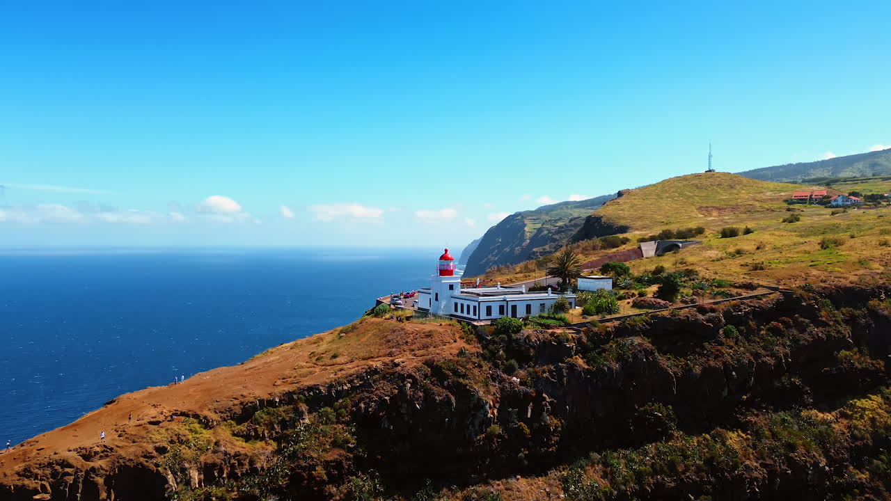 Approaching a beautiful white lighthouse located on the steep mountain on the coast of the Madeira islands. Tourists stand at the observation deck looking at immense waterscape of the North Atlantic Ocean.