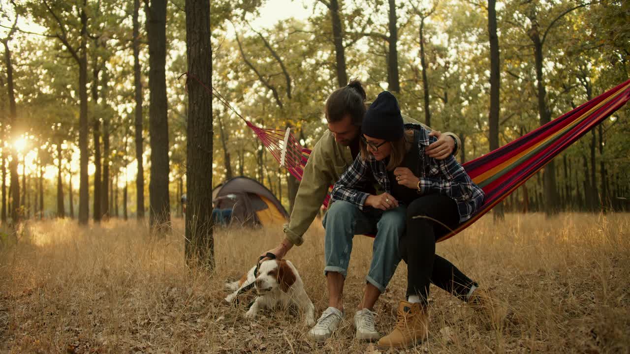 una pareja feliz, un hombre moreno con una chaqueta verde claro y una chica rubia con un sombrero negro, están sentados en una hamaca roja y acarician a su perro marrón blanco contra el fondo de una tienda de campaña gris en un bosque verde de verano soleado