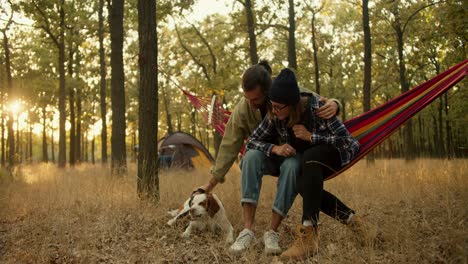 a happy couple, a brunette man in a light green jacket and a blonde girl in a black hat, are sitting on a red hammock and petting their white-brown dog against the background of a gray tent in a sunny summer green forest