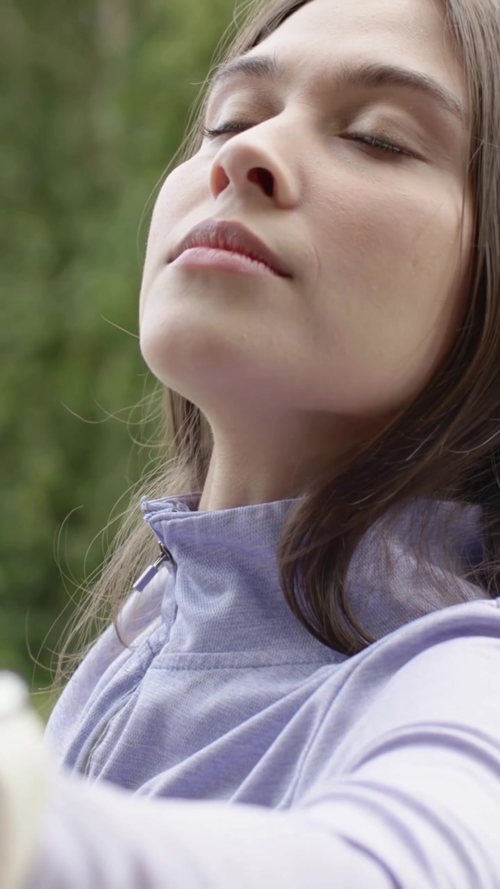 Video verticale: Donna che si tiene il braccio, stringe le labbra, respira e chiude gli occhi, calmandosi al parco, con un top lilla.