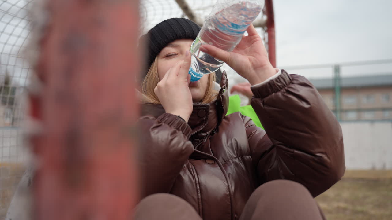 Mujer caucásica bebiendo de una botella grande de plástico sentada junto a una portería, entorno deportivo dinámico al aire libre con cielo nublado, chaqueta abrigada y gorro, recuperación concentrada durante un entrenamiento frío