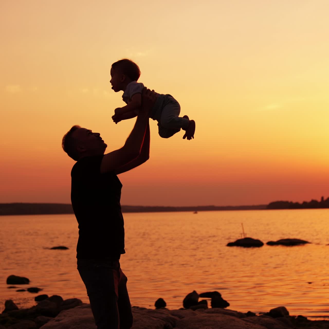 Un padre sorridente lancia in aria il suo bambino. Papà e figlio si godono del tempo in famiglia all'aperto nella natura. Fiume al tramonto sullo sfondo