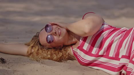 a beautiful young woman wearing sunglasses lies in the white sand and joyfully smiles at the camera on a warm sunny summer day