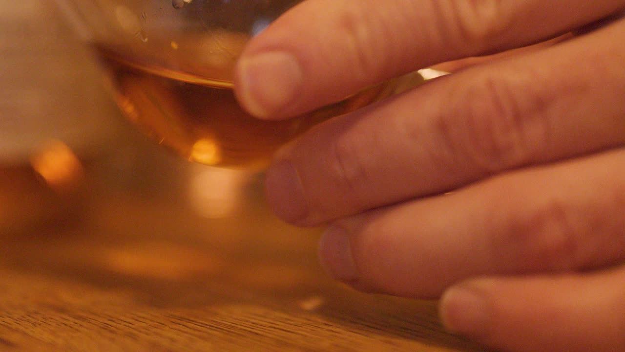 Hand lifts whisky glass from wooden bar under warm, ambient lighting in close-up view