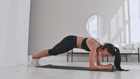 female brunette fitness trainer demonstrating plank knee to chest technique on a mat in home