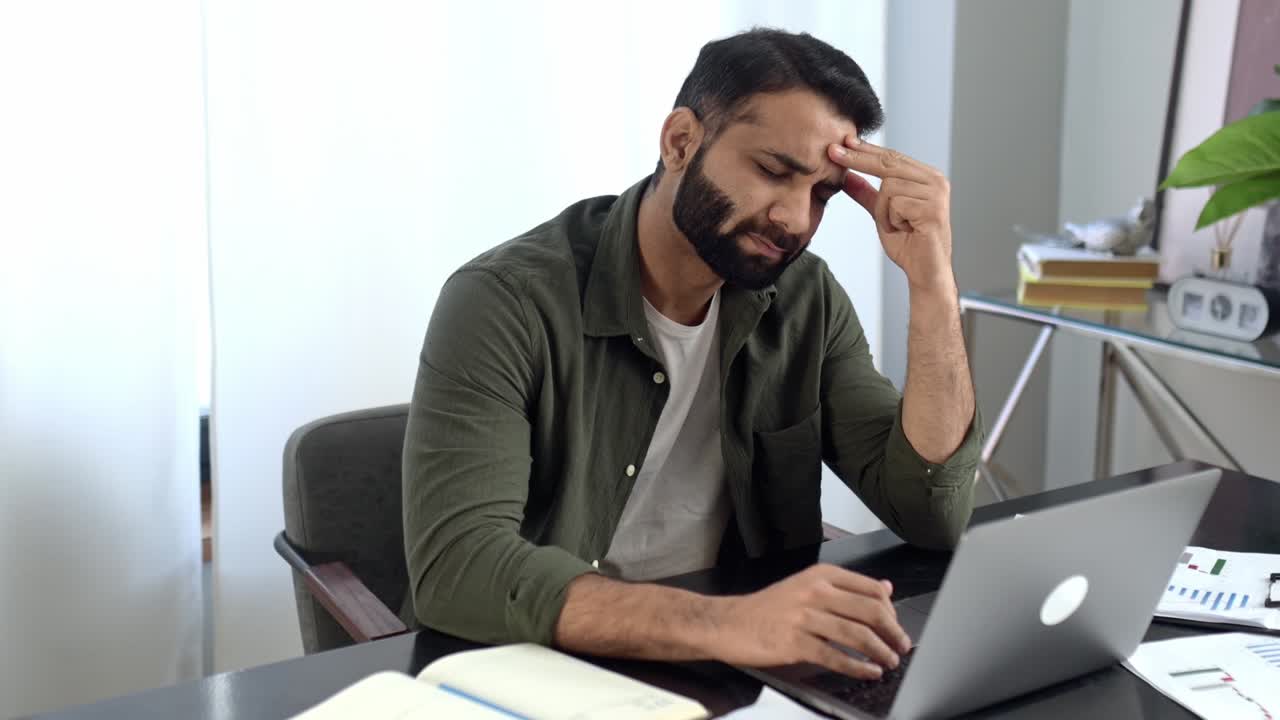 Pensive indian or arabian man in casual clothes, freelancer, designer, stock broker, sitting in modern office, uses a laptop, looking away, thinking about a new project, tired of online work