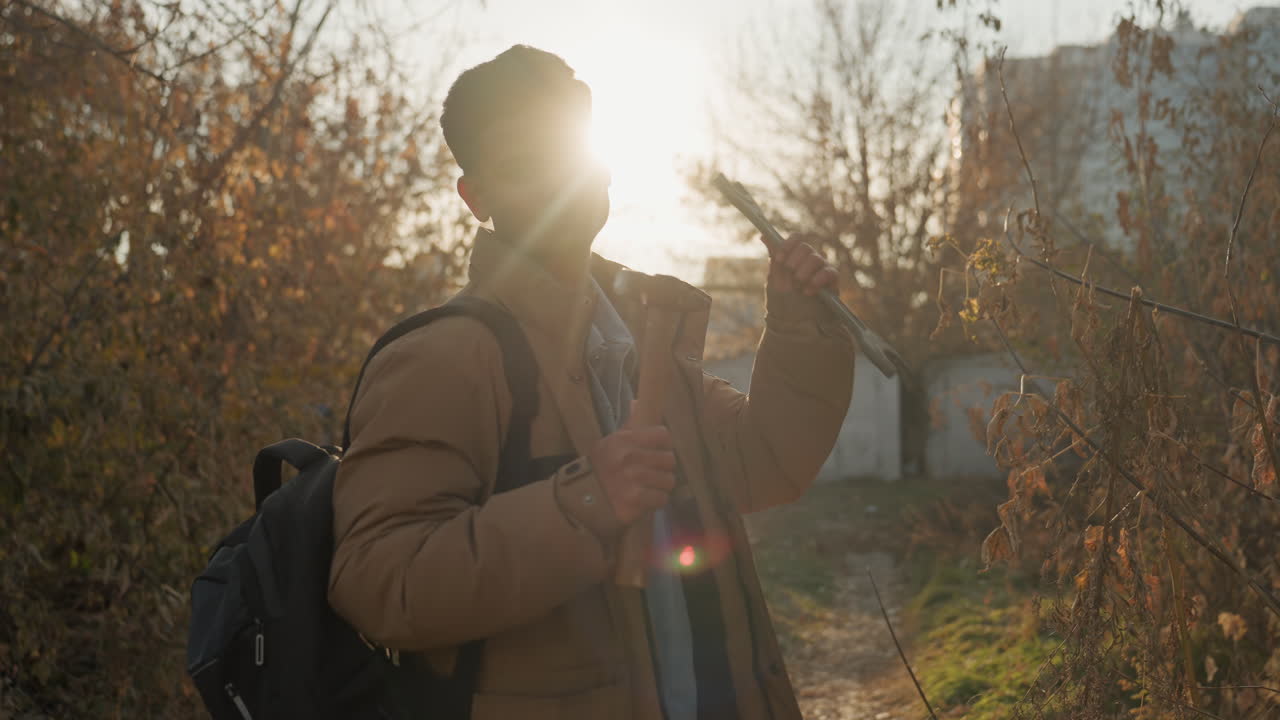 Figurative silhouette on overgrown trail with tools. Contemplative figure holding tools on urban fringe at sunset. Silhouetted South Asian man with pliers and hammer on overrun path during sunset