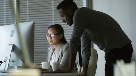 Hombre-Negro-Con-Anteojos-Y-Mujer-Sentada-En-El-Escritorio-De-La-Oficina-Y-Viendo-Algo-En-La-Computadora-Cuando-Trabaja-Tarde-En-La-Noche