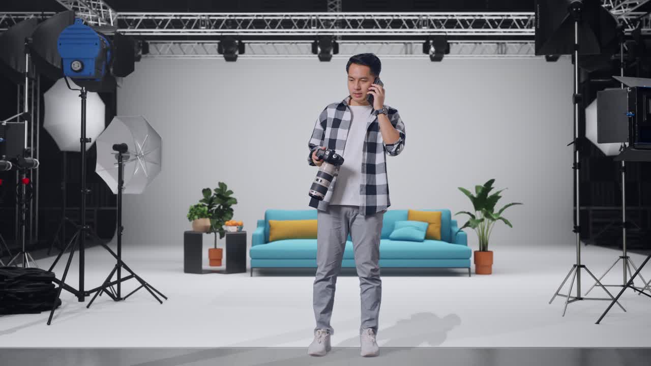 Full Body Of Asian Photographer Holding A Camera In His Hands And Talking On Smartphone While Standing In The Studio