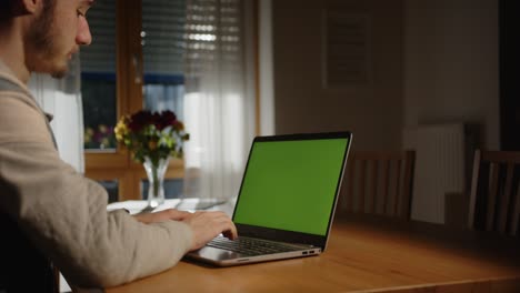 Alguien-Está-Escribiendo-En-La-Computadora-Portátil-Con-Una-Pantalla-Verde-En-La-Sala-De-Estar,-Con-Luz-Del-Exterior