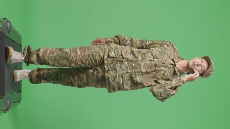 cuerpo lleno de soldado hombre asiático hablando en el teléfono inteligente y mirando a su alrededor mientras camina en el estudio de fondo de la pantalla verde