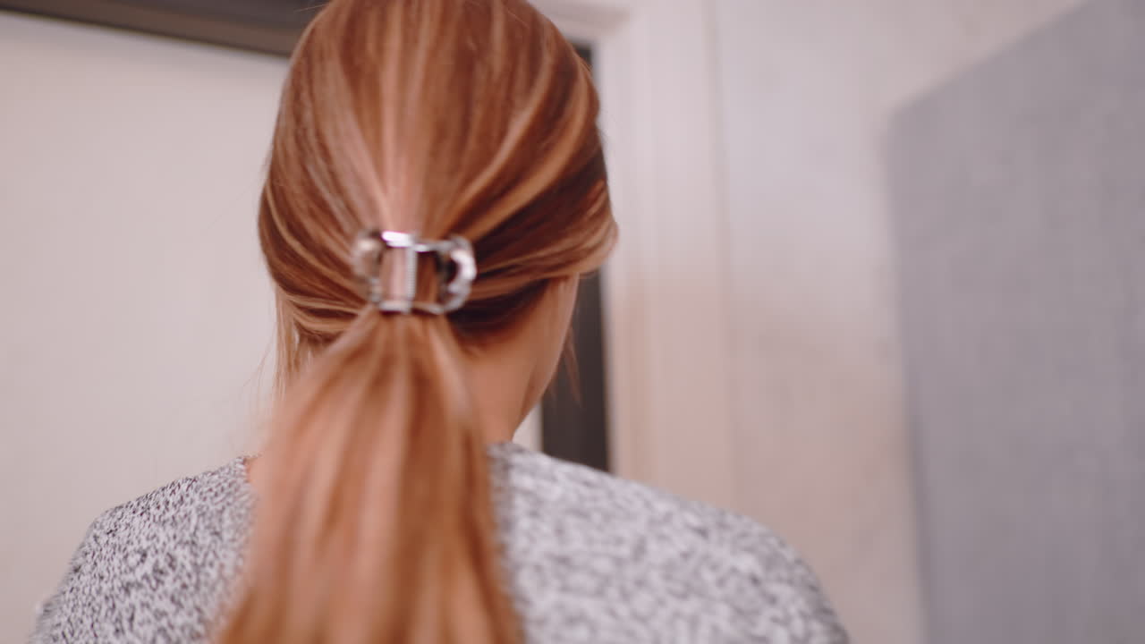Close up of young woman holding black coat as she prepares to leave home, serious expression on her face, standing near wooden coat hanger and wardrobe in softly lit interior hallway