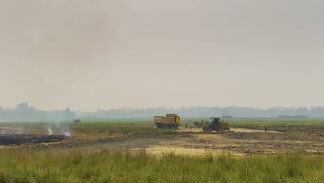 Bulldozer-excavating-earth-removal-next-to-smoking-ash-construction-site,-Bangladesh