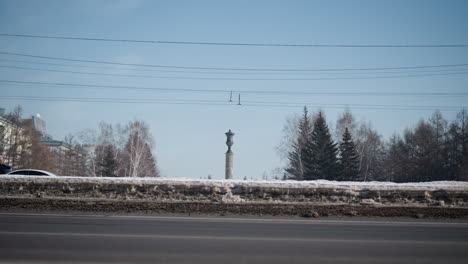 lower view shows busy city road with cars moving on both sides under electric wires, snow piled along curb, winter sun and blue sky over trees and buildings, steady commute scene with motion