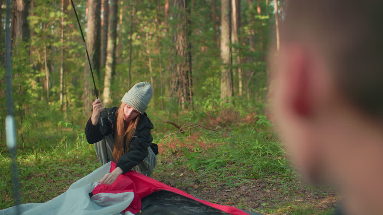 Blurred head appears in foreground observing young girl squatting and fixing tent fabric hook on flexible pole during camping setup in forest