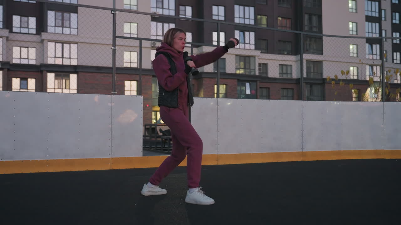 Female participant boxing dumbbells punching air on black asphalt court next to white barrier topped with chain link fence under urban dusk sky wearing maroon hoodie and white sneakers