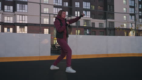 female participant boxing dumbbells punching air on black asphalt court next to white barrier topped with chain link fence under urban dusk sky wearing maroon hoodie and white sneakers
