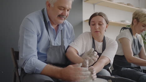 Medium-shot-of-middle-aged-ceramic-artist-teaching-group-elderly-Caucasian-woman-and-senior-man-how-to-wedge-clay-sitting-at-desk-in-art-studio.-People-enjoying-talking-at-work