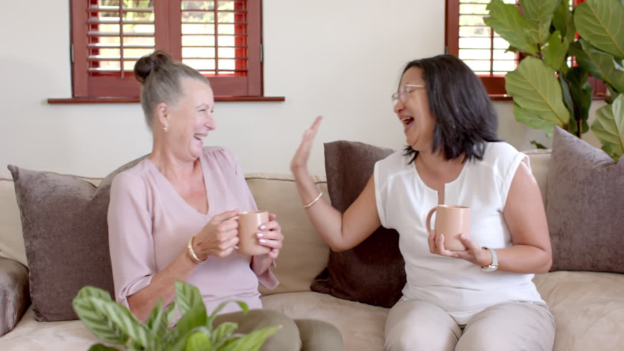 Senior diverse women laughing together on sofa while enjoying coffee at home
