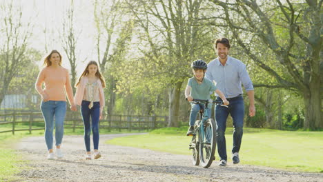 family on summer walk in countryside with father teaching son to ride bike