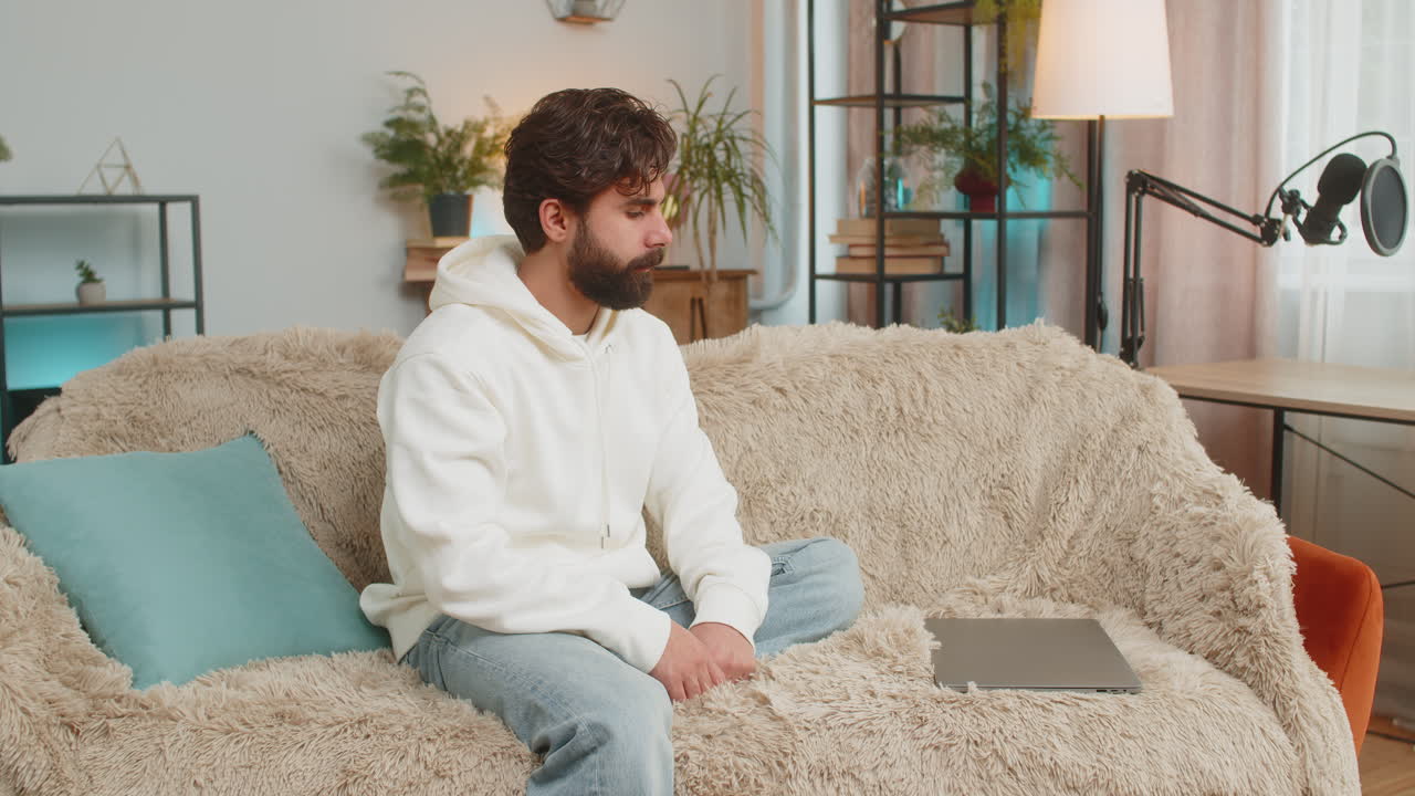 Young indian man sitting on sofa couch opens laptop computer start working in living room apartment