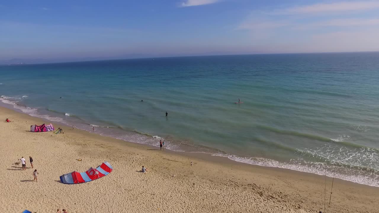 Paning view of the coast. Waves crashing on the beach line, some fishing rods, a couple unmbrellas and a kite surf seen on the sand.