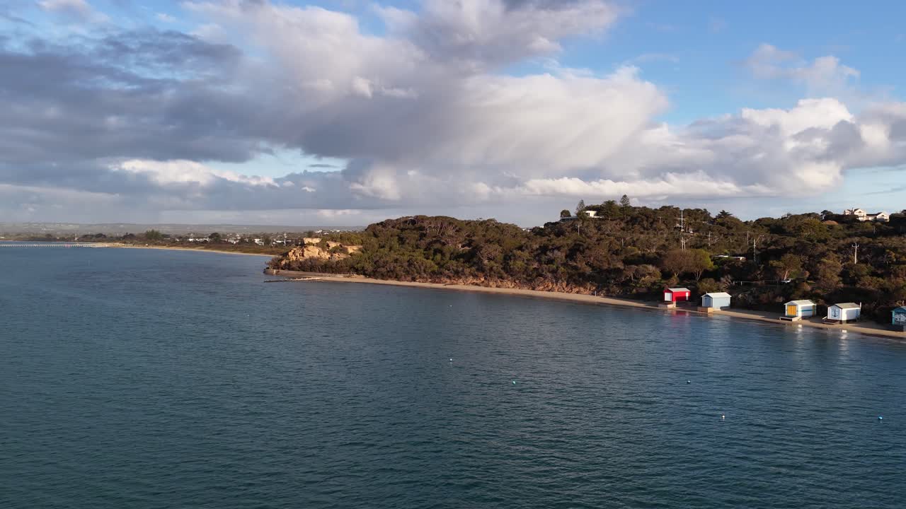 Drone glides above calm bay, revealing vibrant beach huts, natural shoreline, and dramatic afternoon clouds