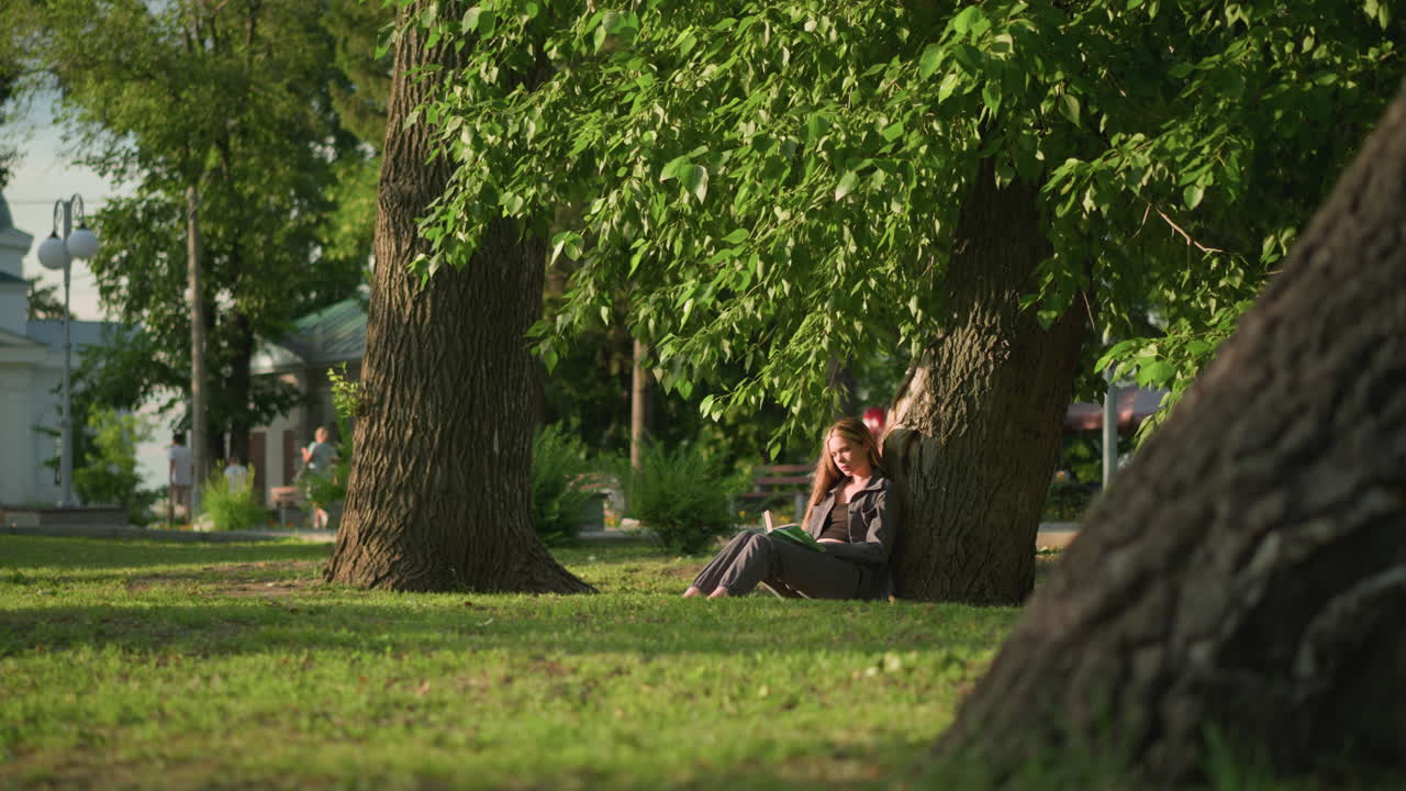 야외에 앉아있는 여인, 잔디에 있는 나무에 기대어, 아래에서 책을 읽고, 나무 잎이 바람에 부드럽게 흔들리고, 배경은 건물과 사람들이 걸어가는 것을 보여줍니다.
